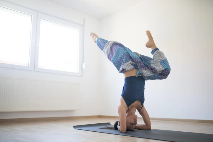 Junge Frau trägt eine Haremshose und macht auf einer dünnen Sportmatte einen Kopfstand. 