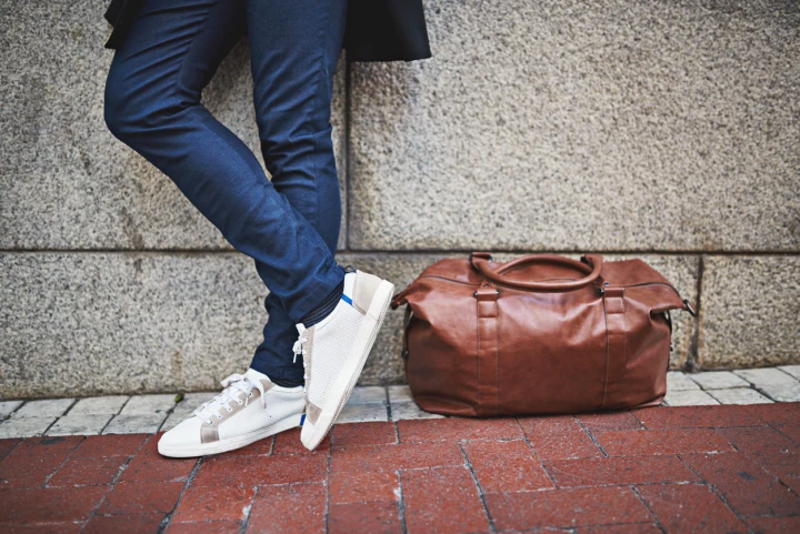 Mann in Jeans und Sneakern lehnt mit seinem braunen Weekender gegen eine Steinwand